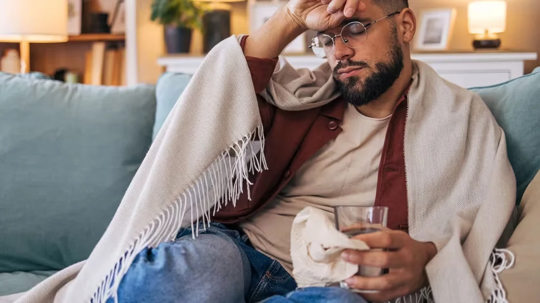 Homem sentado no sofá, enrolado em um cobertor, com expressão de cansaço e segurando um copo de água e um lenço, ilustrando sintomas comuns usados para explicar a diferença entre gripe e resfriado, como mal-estar, fadiga e indisposição.