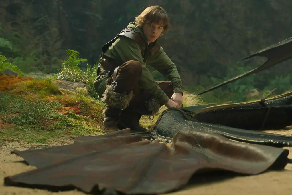 Cena da versão live-action de Como Treinar Seu Dragão mostrando Soluço ajoelhado ao lado de Banguela na floresta, destacando o realismo do dragão e as diferenças visuais em relação à animação original. Crédito: Universal Pictures