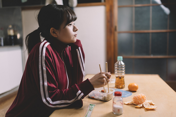 Estudante japonesa do ensino médio em casa, usando uniforme esportivo, fazendo refeição simples à mesa — imagem que representa a rotina real e mostra como o ensino médio nos animes é diferente do ensino médio real.