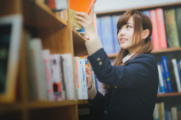 Estudante japonesa do ensino médio escolhendo livro na biblioteca escolar — imagem que representa a rotina acadêmica e ilustra como o ensino médio nos animes é diferente do ensino médio real.