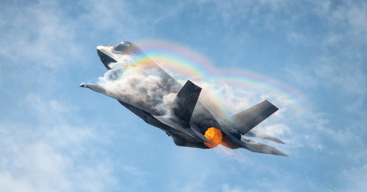 Avião de caça F-35 em alta velocidade com pós-combustão acesa, envolto por nuvens de condensação e arco-íris atmosférico no céu azul durante manobra aérea.